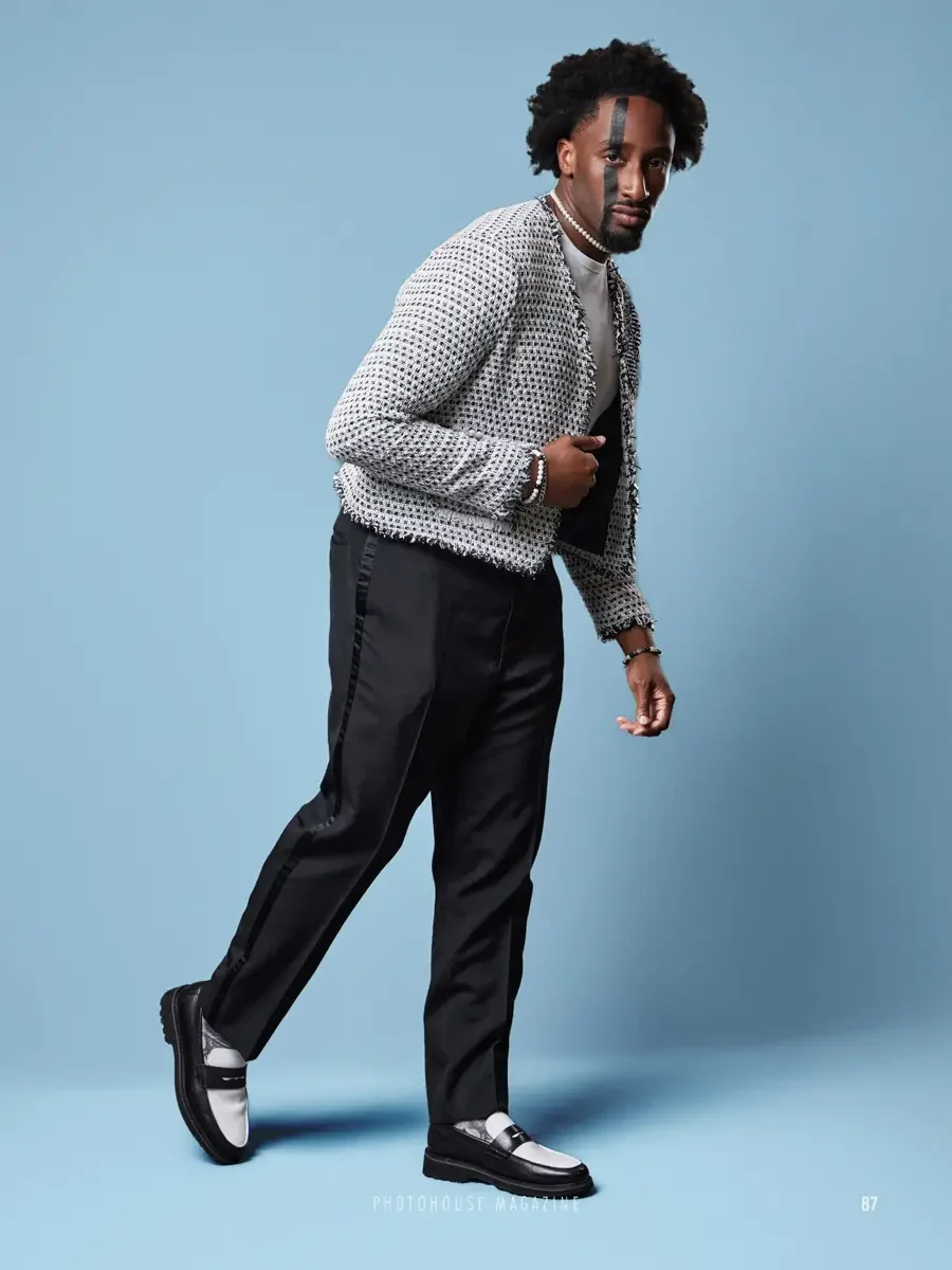 JUS10H 2025 Photohouse Magazine feature, model in motion wearing white and black fringe-trimmed boucle cardigan over white turtleneck and black tailored trousers with satin side stripe, two-tone penny loafers, light blue background, photographed by S