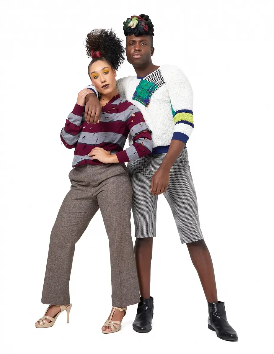 Model portfolio development session, high fashion editorial look, two models standing close, man in white patchwork bouclé knit sweater and houndstooth bermuda shorts with arm around woman in burgundy and heather grey destroyed-knit stripe sweater an