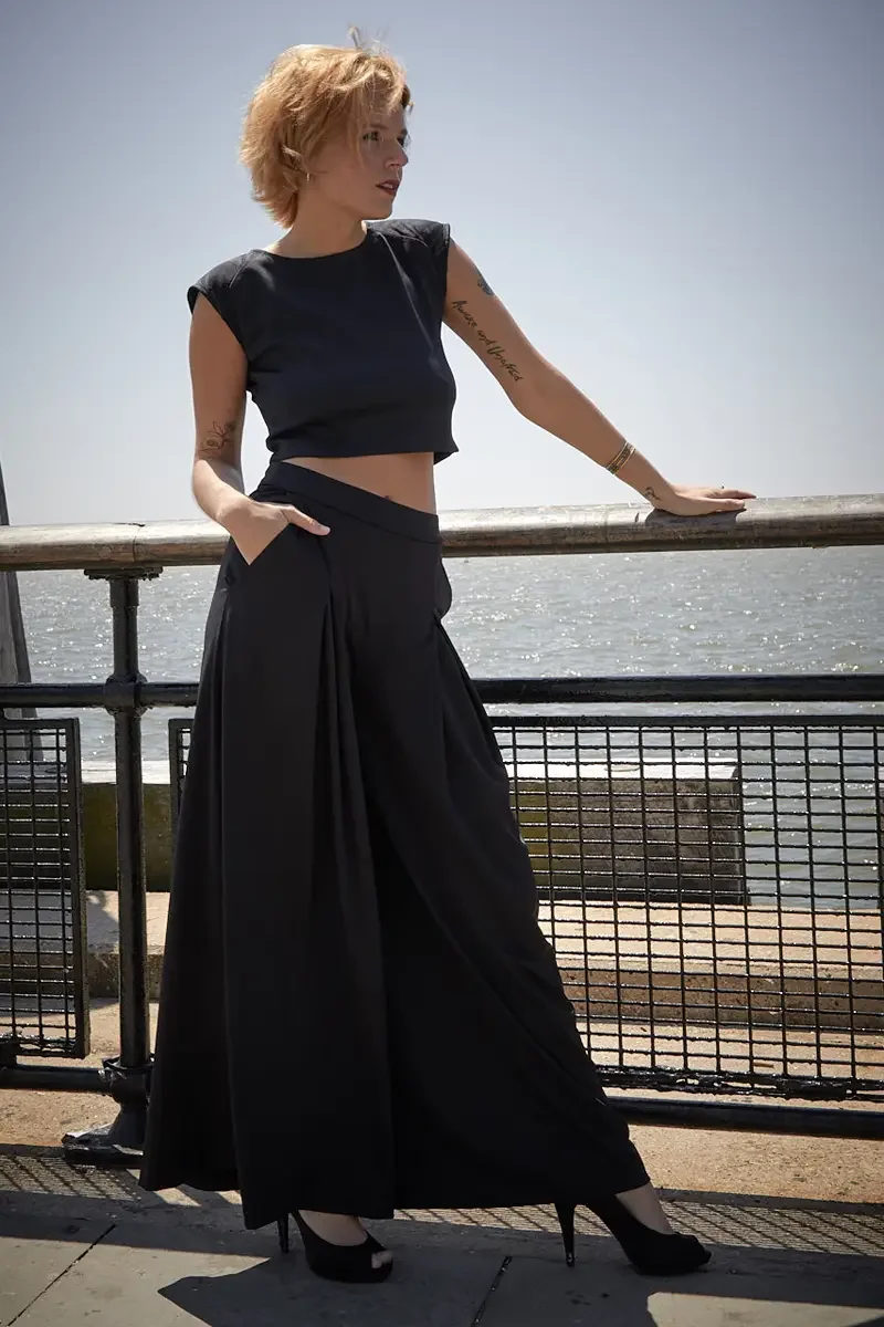 Satine, full length, leaning on waterfront railing, black structured cap-sleeve crop top with shoulder detail, black wide-leg pleated palazzo pants with wide waistband, black platform peep-toe heels, script and bird tattoo on right arm, gold bangle, 