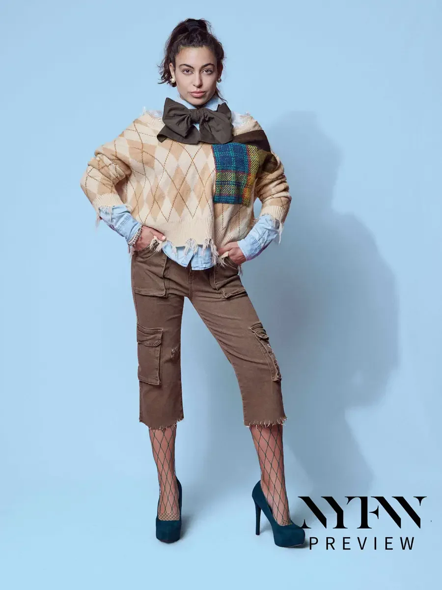 JUS10H 2025 social media, model with hands on hips in camel argyle fringe sweater with multicolor tartan patchwork panel, olive oversized bow, and brown distressed cargo capris over fishnet tights and teal platform heels, light blue background, photo