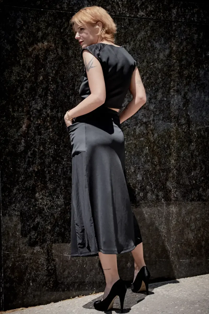 Satine, full length, back to camera, glancing over shoulder, black structured cap-sleeve crop top, charcoal grey A-line midi skirt, black satin peep-toe stiletto heels, gold bangle, bird tattoo on upper right arm visible, granite wall, Battery Park a