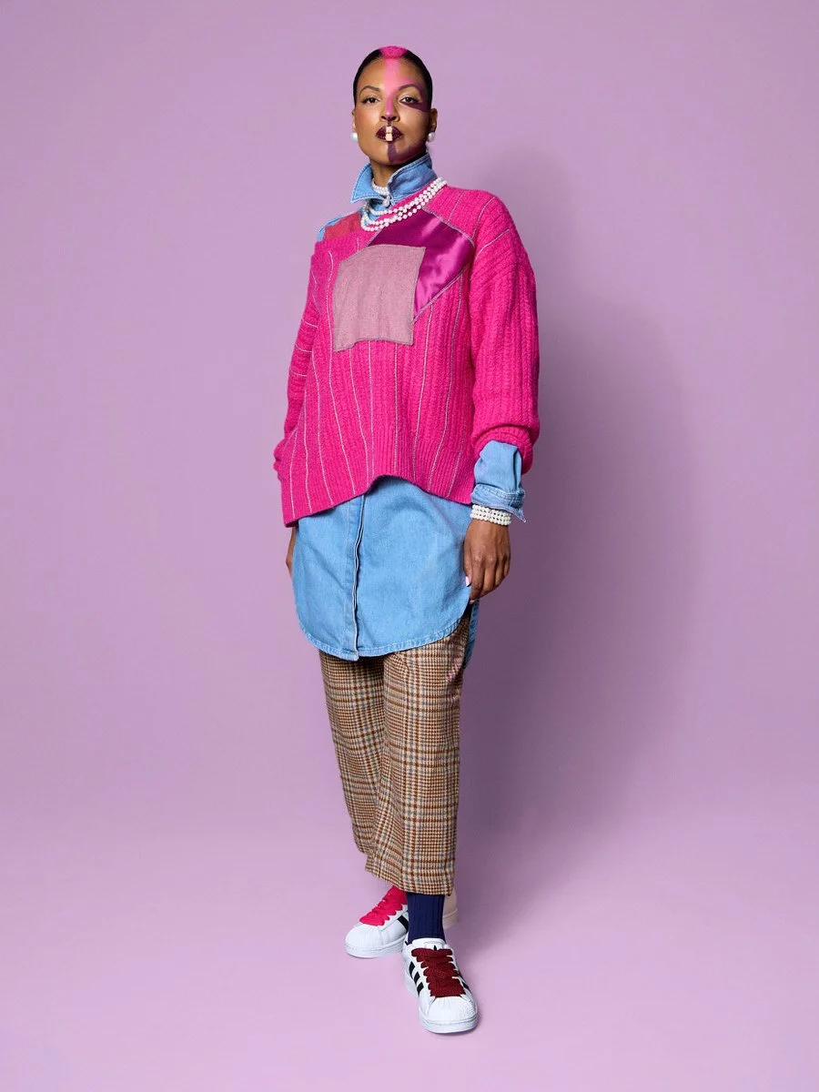 A woman standing against a purple background, wearing a pink and purple patchwork sweater over a blue denim shirt, plaid pants, and sneakers with pink and black laces. She has makeup with bold pink and purple colors and is accessorized with pearl nec