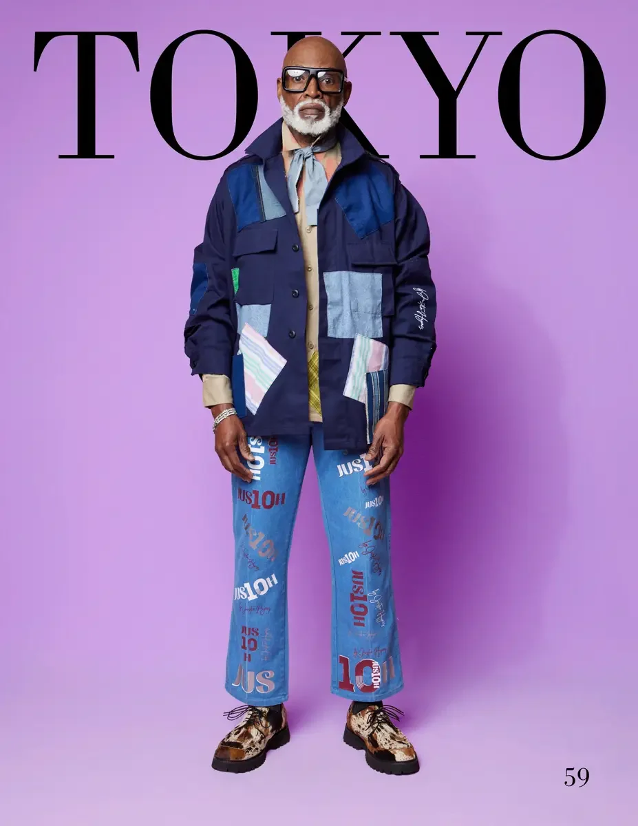 JUS10H 2025 Tokyo lookbook, a male model wearing a navy multi-pocket field jacket with mixed pastel stripe, plaid, denim, and chambray patchwork appliqués and a script logo, over a tan button-down shirt, light blue JUS10H all-over logo-print flared j