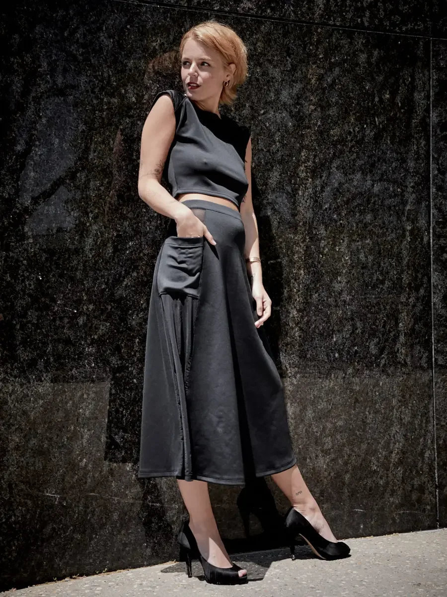 Satine, full length, profile, hand in skirt pocket, black structured cap-sleeve crop top, charcoal grey A-line midi skirt with wide waistband and sheer mesh hip panel, black satin peep-toe stiletto heels, gold bangle, small ankle tattoo visible, gran