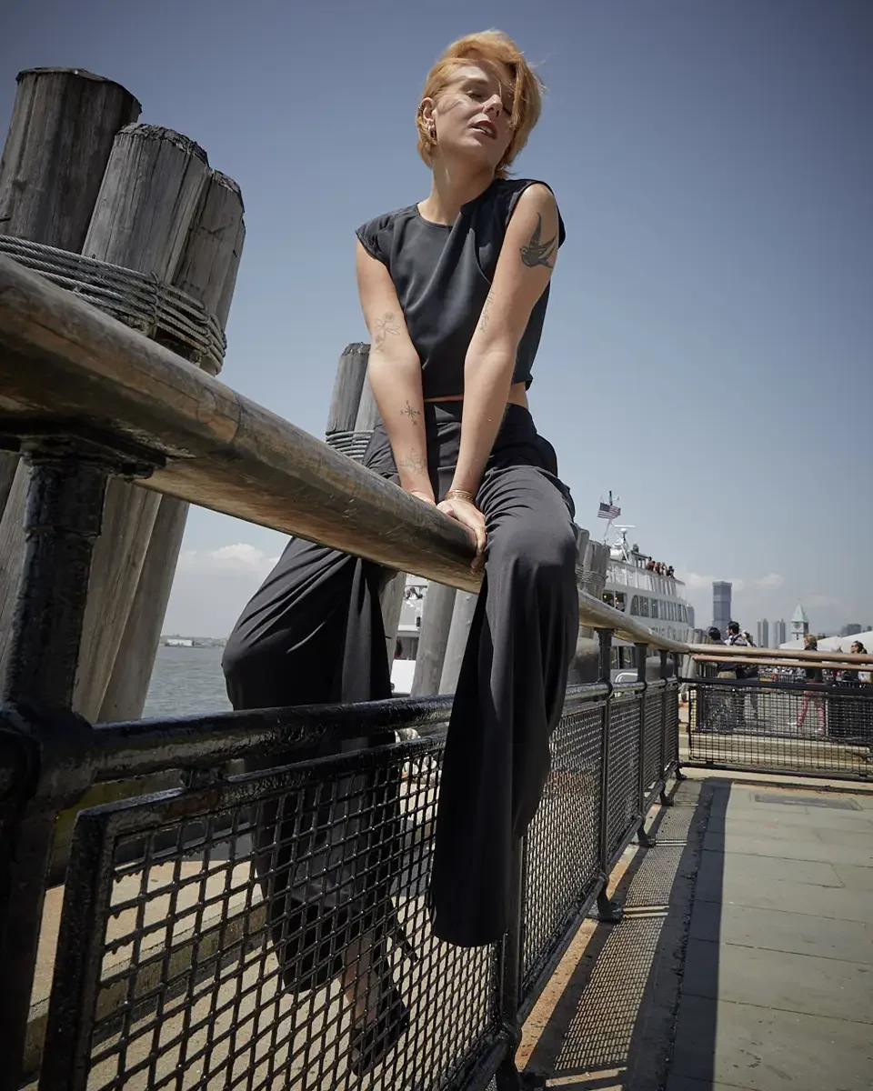 Satine, full length, seated on pier railing, low camera angle, leaning forward with hands clasped, black structured cap-sleeve crop top, black wide-leg pleated palazzo pants, gold bangles, bird tattoo and floral tattoo on arms visible, Battery Park p