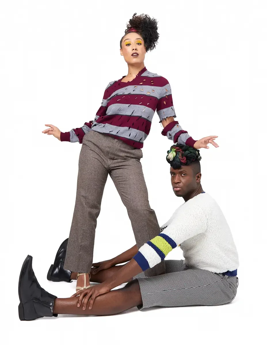 Model portfolio development session, high fashion editorial look, woman standing in burgundy and heather grey destroyed-knit stripe sweater and straight-leg heathered trousers with both arms extended outward, man seated on floor in white patchwork bo