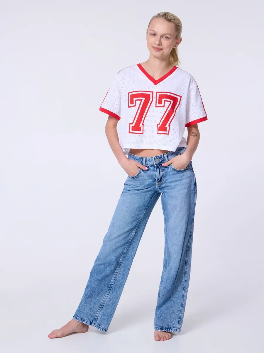 Model portfolio development session, commercial look, white mesh crop football jersey with red number 77 and red trim, low-rise light wash wide-leg jeans, full-length, facing camera, both hands in front pockets, weight shifted to one hip, easy smile,