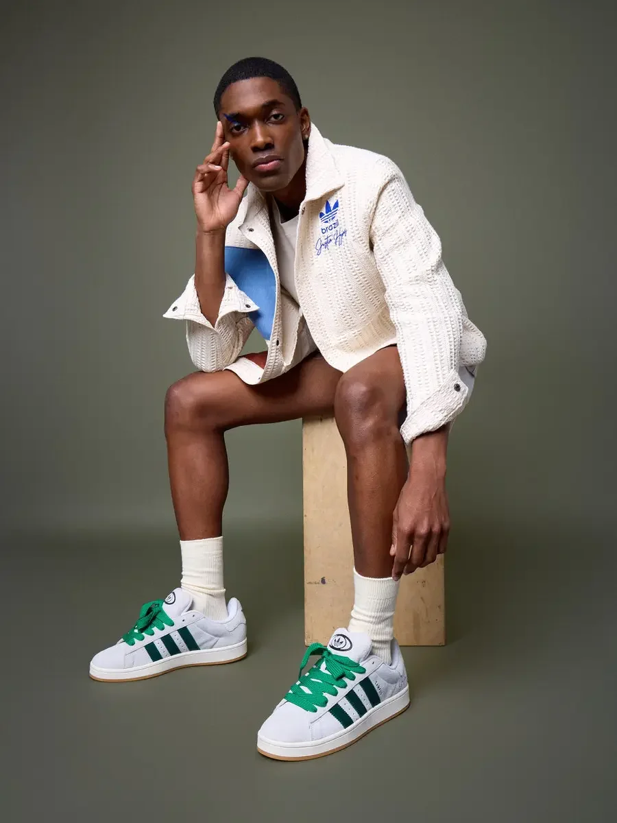 JUS10H x Adidas Brasil campaign, model seated on wood block with hand raised to face, white textured patchwork jacket with blue and terracotta suede panels, Adidas Campus 00s in white with dark green stripes and green laces, sage green background, ph