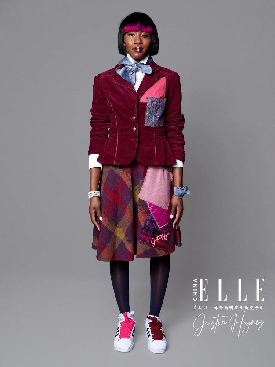 Fashion model wearing a colorful patchwork jacket, plaid skirt, black tights, and sneakers, standing against a gray background.