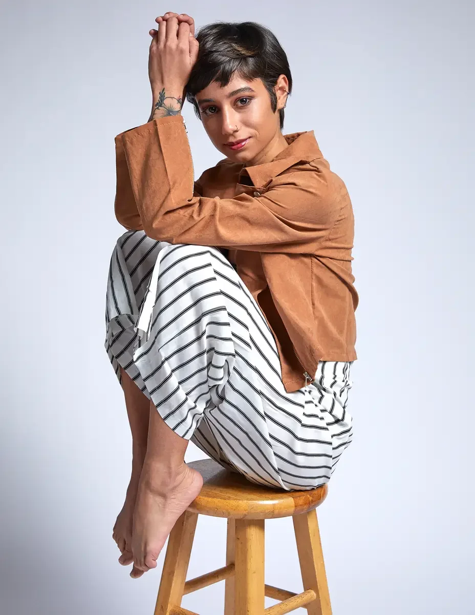 Eloissa, seated, legs tucked, tan suede jacket and striped wide-leg trousers, floral forearm tattoo, studio stool, East Williamsburg studio test shoot, makeup by Schanica Pickens, photographed by Scott Parker Photo