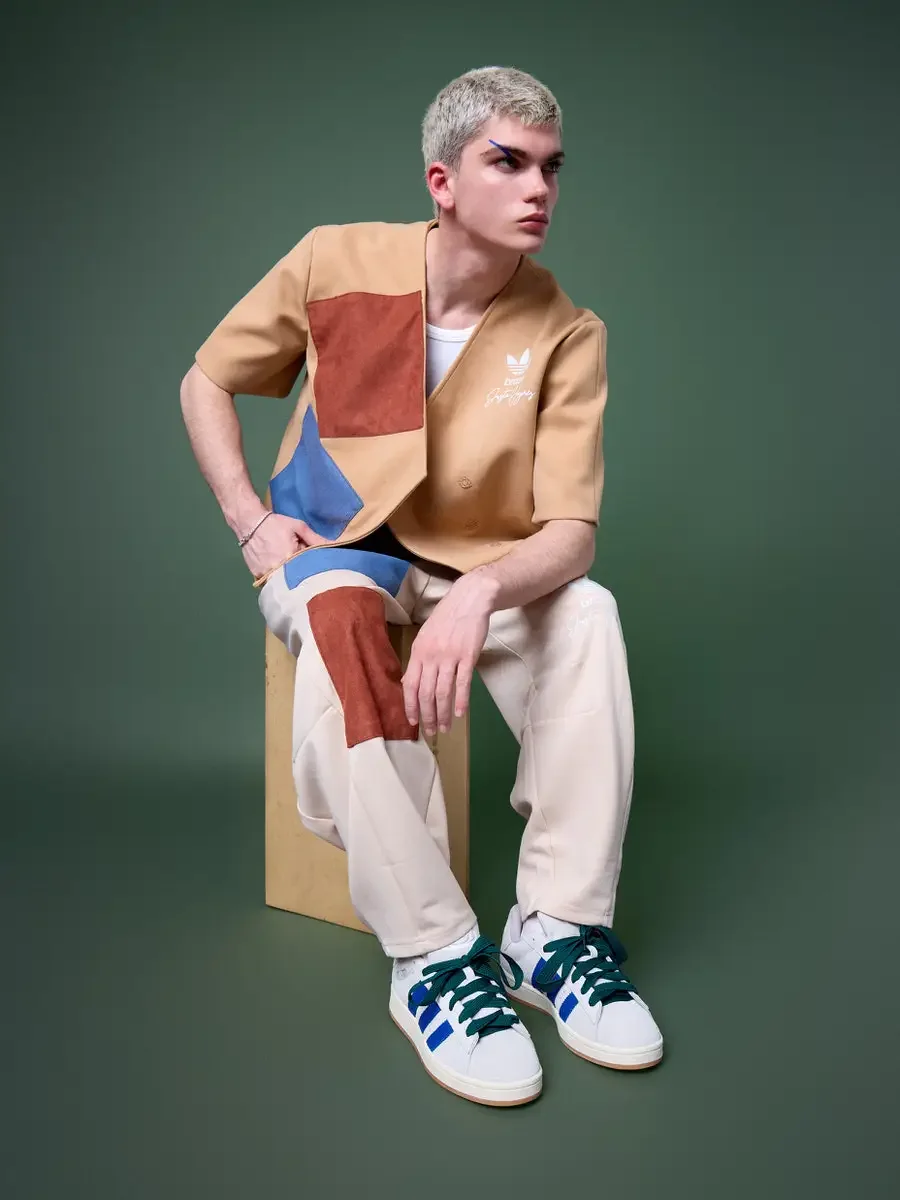 JUS10H x Adidas Brasil campaign, model seated on wood block with hand on hip, tan patchwork short-sleeve vest with terracotta and blue suede panels over cream wide-leg patchwork trousers, Adidas Campus 00s in white with blue stripes and dark green la