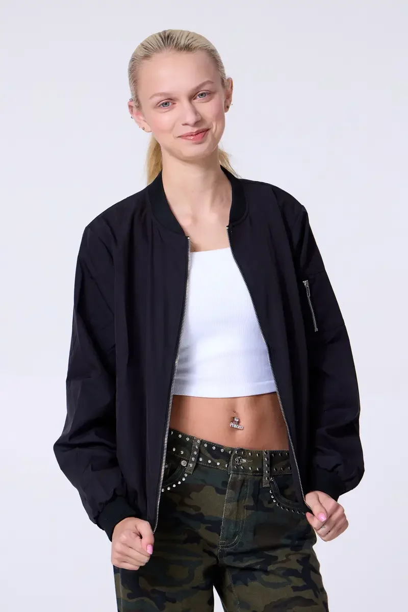 Model portfolio development session, commercial look, black bomber jacket open over white crop tank, low-rise studded camouflage wide-leg trousers with silver stud waistband detail, three-quarter, facing camera, hands at lower jacket hem, relaxed smi