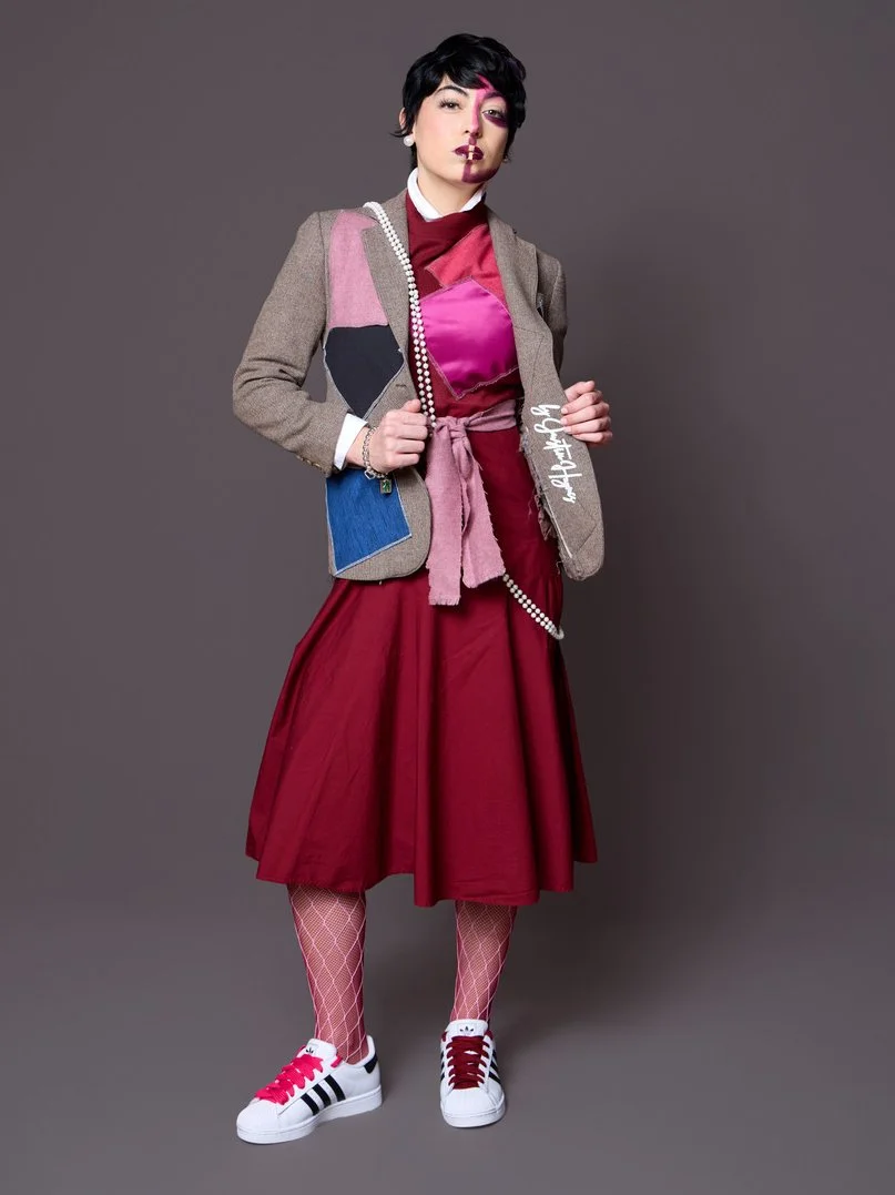 Young woman with short black hair and pink streaks, wearing bold makeup, a colorful patchwork blazer, a shiny burgundy top, a long red skirt, pink fishnet stockings, and white sneakers with black and pink details, standing against a plain grey backgr