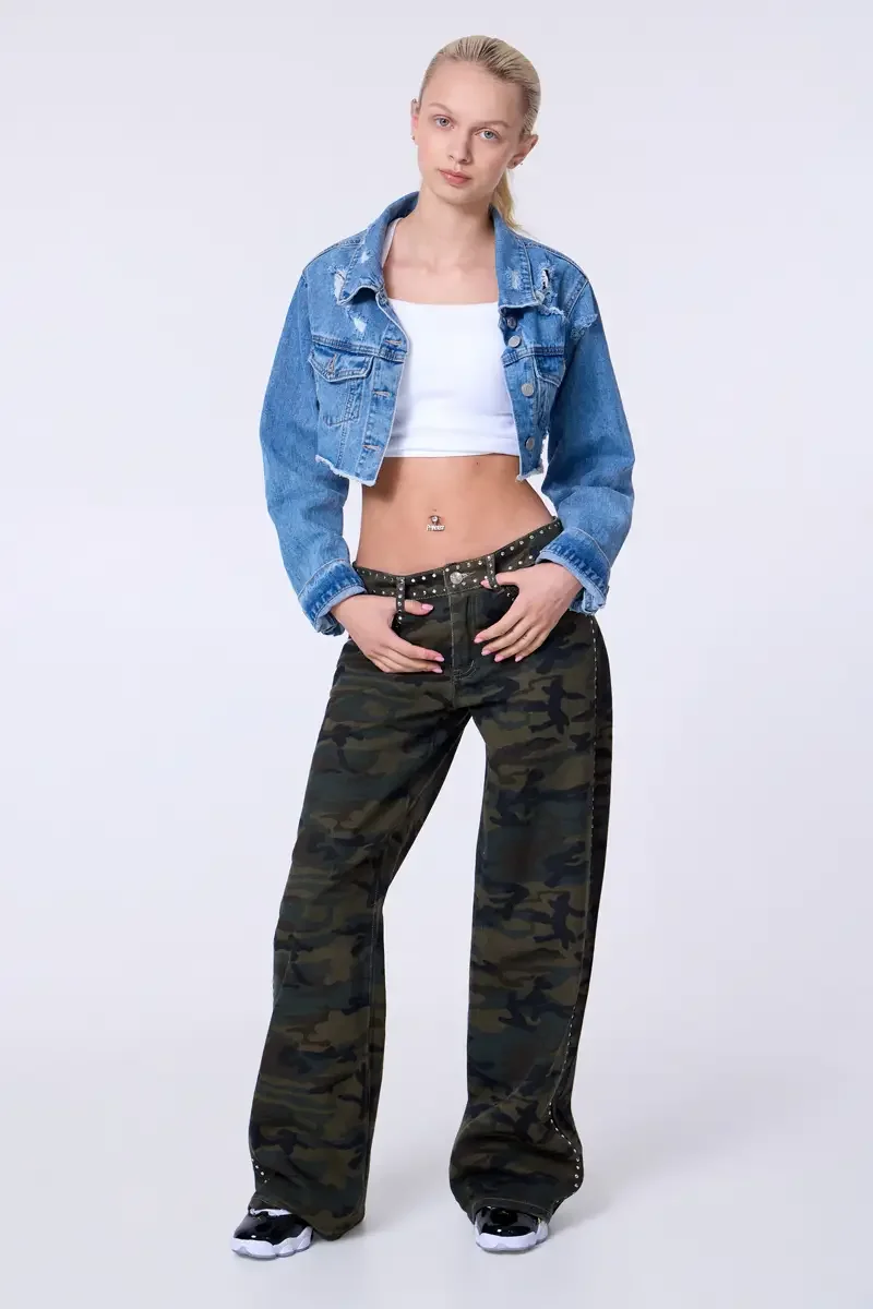 Model portfolio development session, commercial look, cropped distressed blue denim jacket over white crop tank, low-rise studded camouflage wide-leg trousers with silver stud trim at waistband and side seam, full-length, facing camera, both hands at