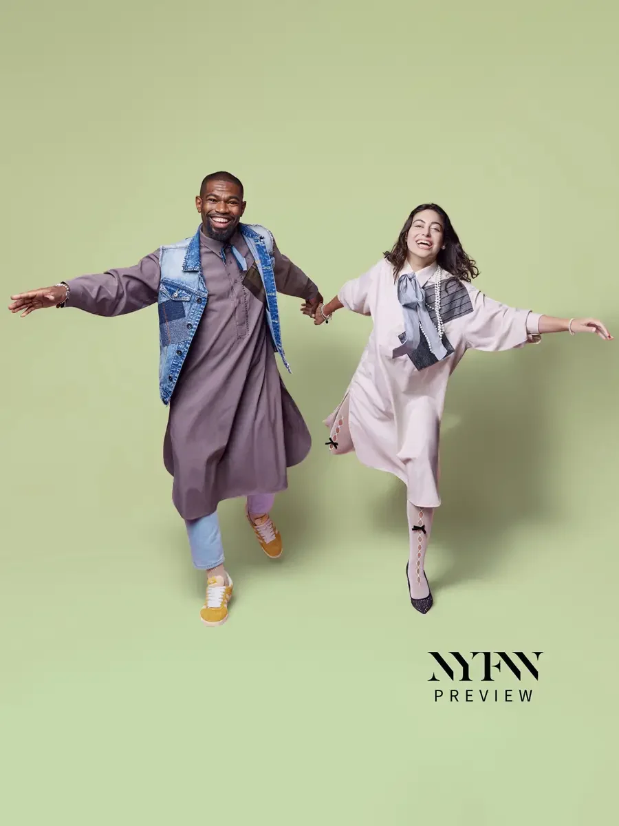 JUS10H 2025 billboard Times Square, two models jumping and holding hands, male in mauve longline shirt-dress with patchwork denim vest over light blue trousers and yellow sneakers, female in blush oversized shirt-dress with striped bow and pearl neck
