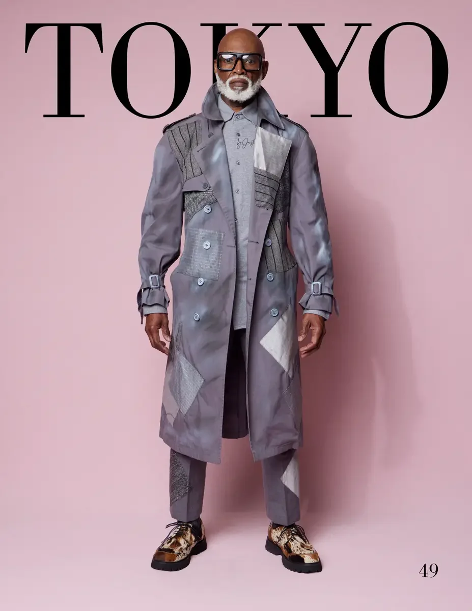 JUS10H 2025 Tokyo lookbook, a male model wearing a gray double-breasted trench coat with mixed patchwork appliqués including striped, houndstooth, crocodile-texture, and geometric panels, over a gray button-down shirt with script embroidery and match