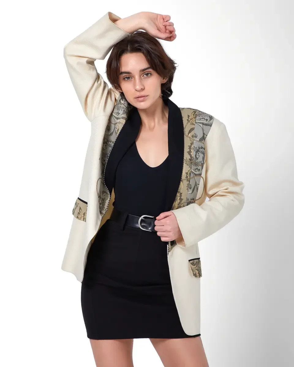 Tess, three-quarter crop, one arm raised overhead, cream textured blazer with baroque tapestry lapel and pocket trim, black deep-V bodysuit, black fitted mini skirt, wide black leather belt with silver D-ring buckle, New York studio test shoot, makeu