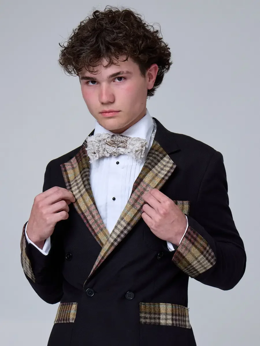 JUS10H 2025 New York Bridal Fashion Week, black double-breasted suit with plaid lapels, pocket trim, and cuffs, styled with a dimensional lace bow tie and pleated white dress shirt, photographed by Scott Parker Photo