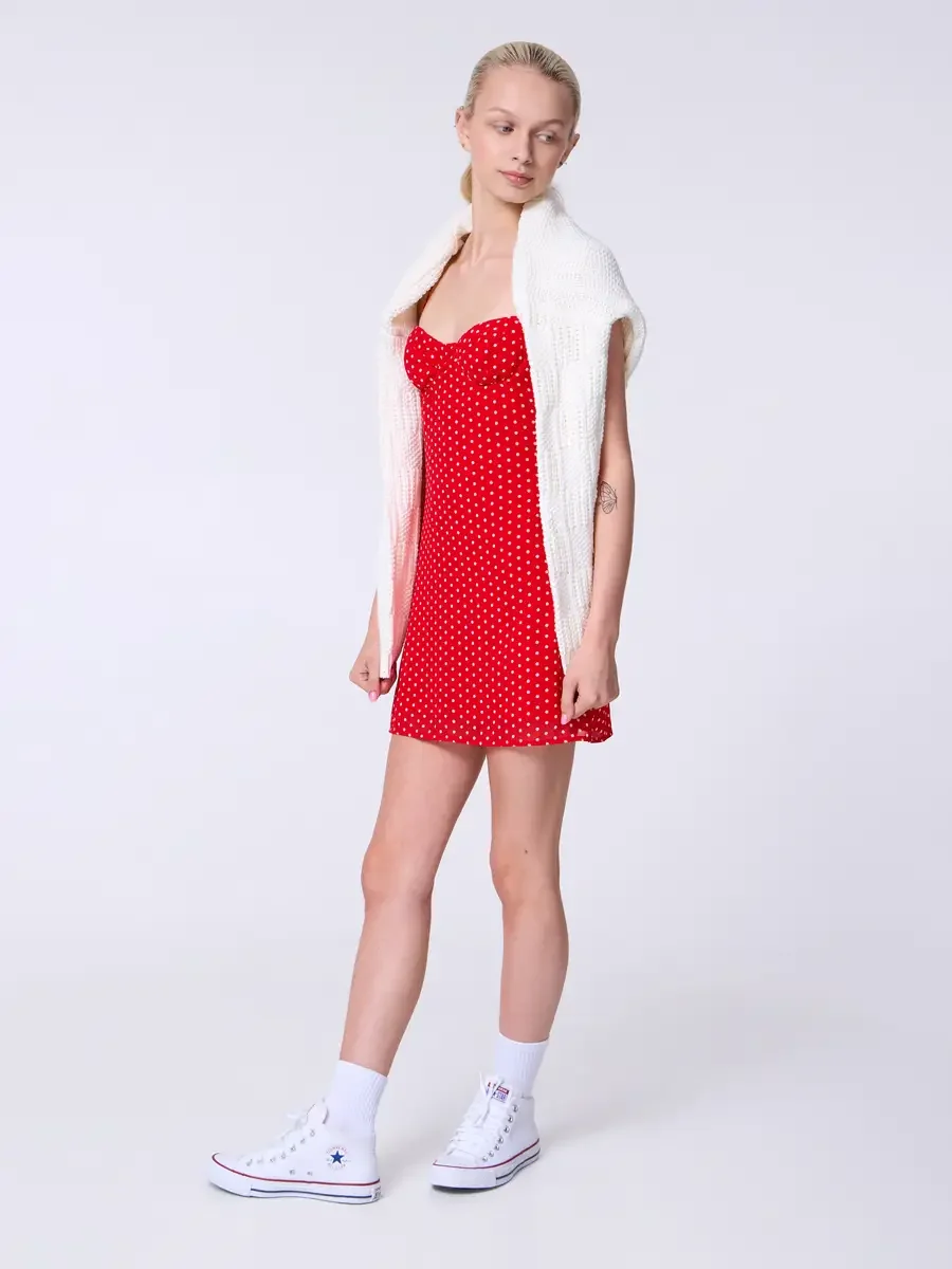 Model portfolio development session, commercial look, red polka dot sweetheart mini dress with cream chunky knit cardigan draped over shoulders as accessory, full-length, slight turn, gaze downward, cardigan worn loose off both shoulders, white high-