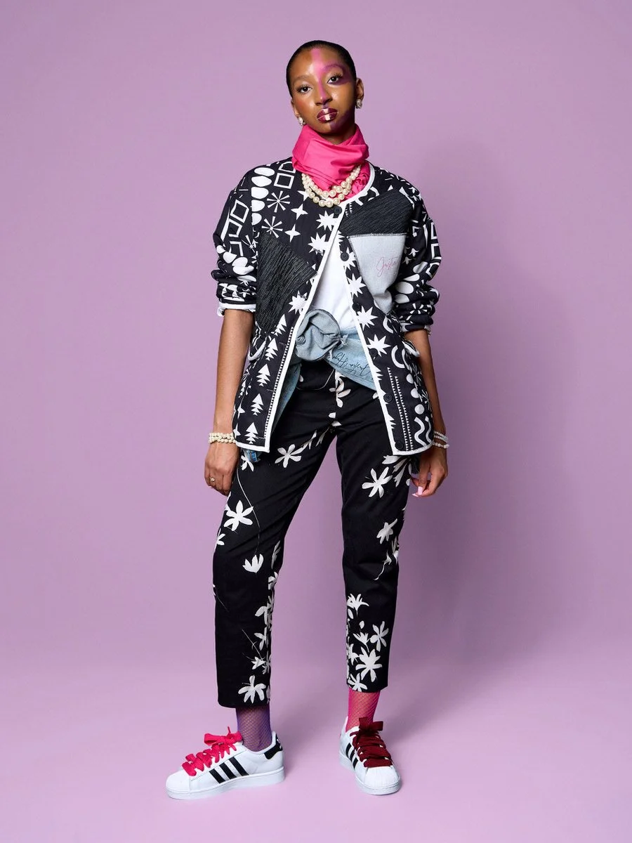 Woman wearing a black and white floral and geometric patterned jacket and pants, pink neck scarf, pearl jewelry, white sneakers with black and red stripes, standing against a purple background.
