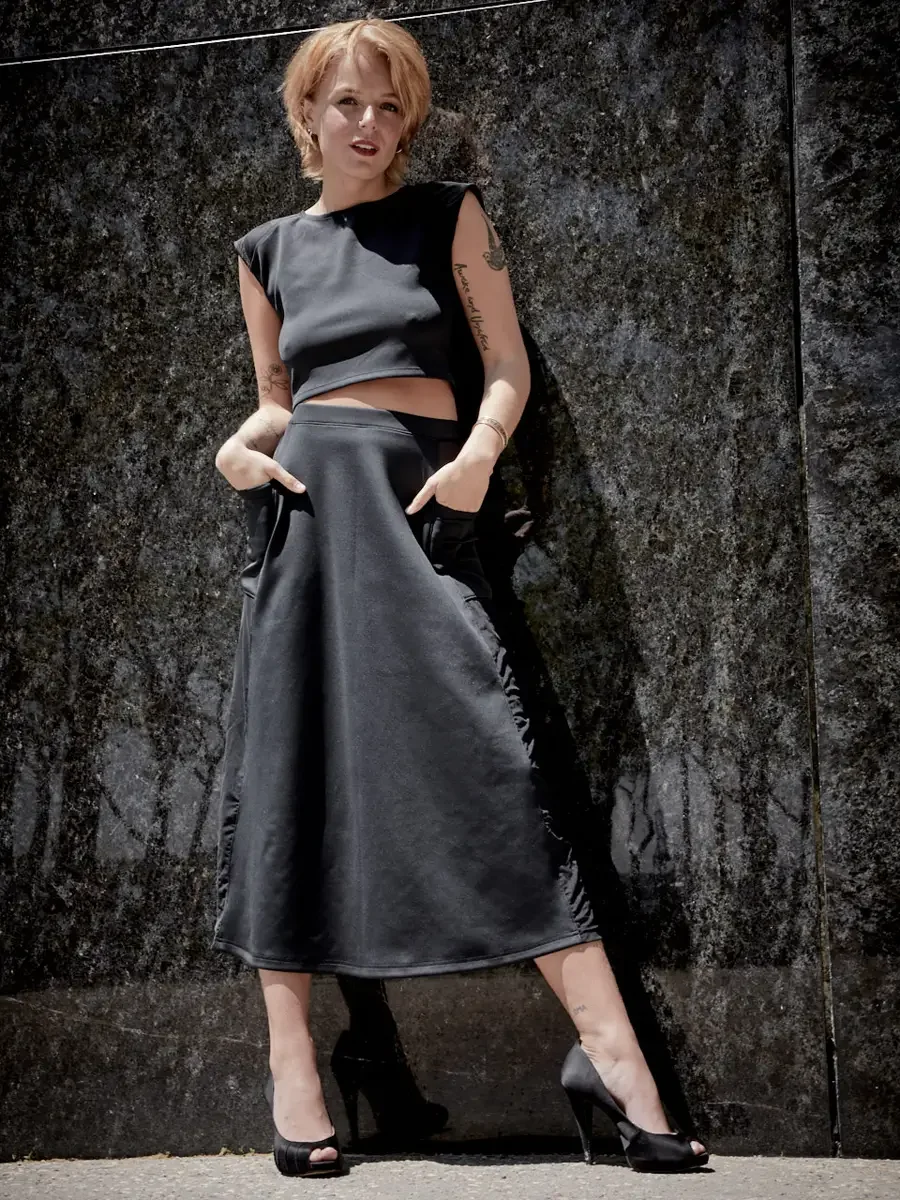 Satine, full length, both hands in skirt pockets, black structured cap-sleeve crop top, charcoal grey A-line midi skirt with wide waistband and sheer mesh hip panel, black satin peep-toe stiletto heels, script tattoo on right arm visible, granite wal