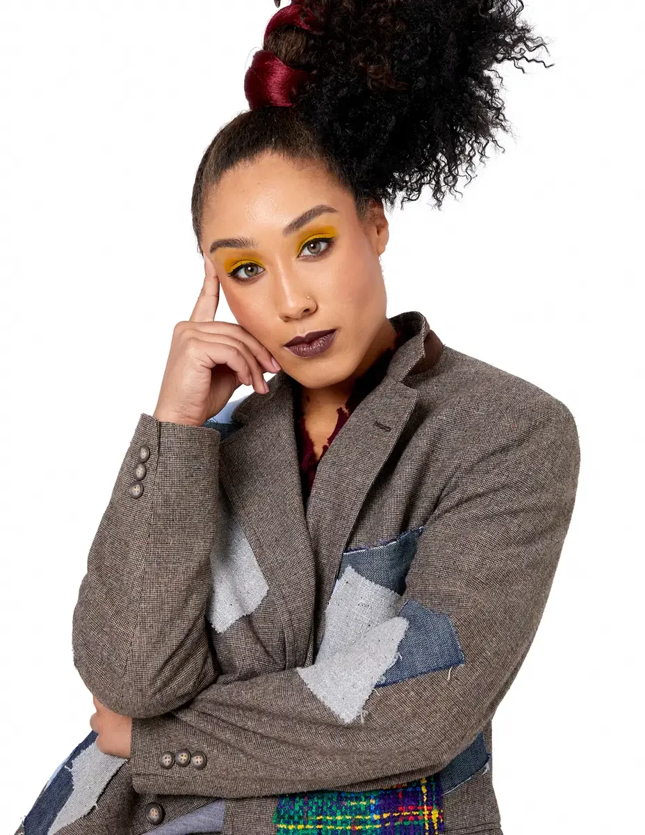 Model portfolio development session, high fashion editorial look, oversized heathered blazer with mixed fabric appliqué panels, three-quarter, arms crossed, one finger raised to temple, contemplative expression, gold editorial eye makeup, dark lip, w
