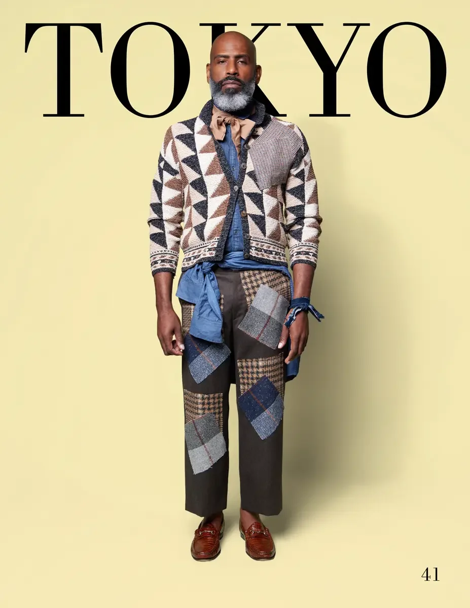 JUS10H 2025 Tokyo lookbook, a male model wearing a cream, brown, and black geometric patterned knit cardigan over a chambray denim shirt tied at the waist with a blue denim sash, dark olive trousers with mixed plaid and tweed patchwork appliqués, a p
