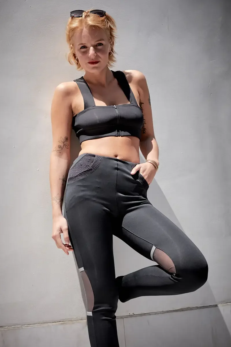 Satine, full length, leaning against concrete wall, one knee raised, black zip-front structured crop top, black high-waist leggings with geometric embossed pocket, sheer mesh panel inserts and reflective white stripe, sunglasses on head, gold bangles