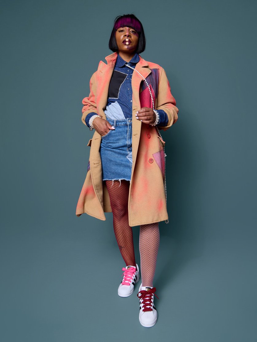 A woman wearing a colorful coat, denim shirt, denim skirt, fishnet tights, and sneakers with pink and red laces, standing against a plain background.