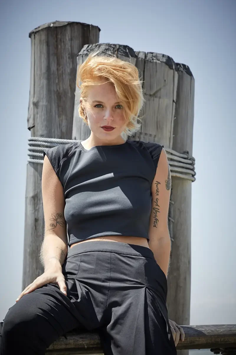Satine, close-up, seated against wooden pier piling, black structured cap-sleeve crop top, black wide-leg palazzo pants, script tattoo reading Awake and Unafraid on right upper arm, floral tattoo on left forearm, nose ring, Battery Park pier test sho