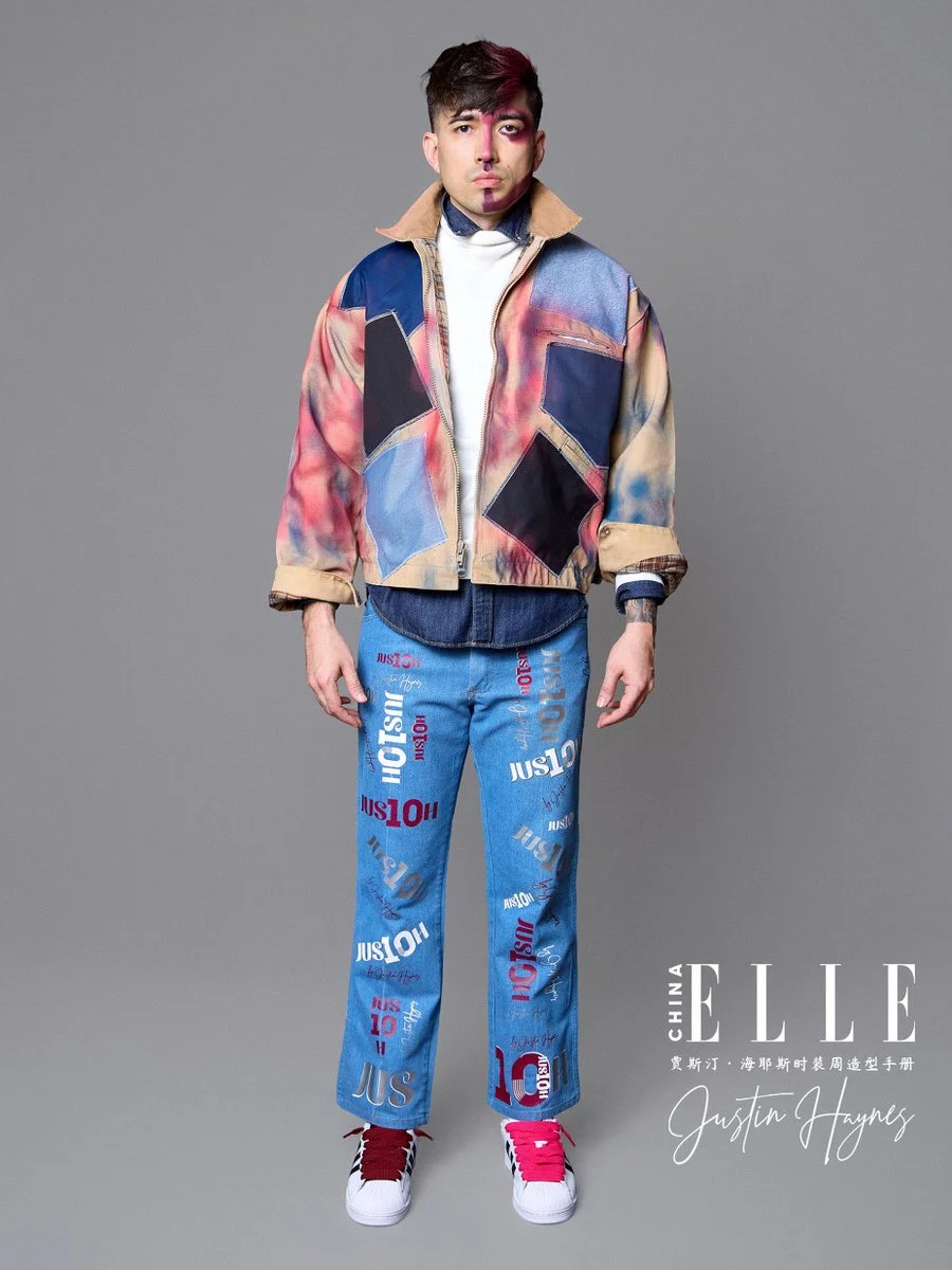 Young man in colorful, graphic-patterned jacket and blue jeans with text and logo print, standing against a plain gray background.