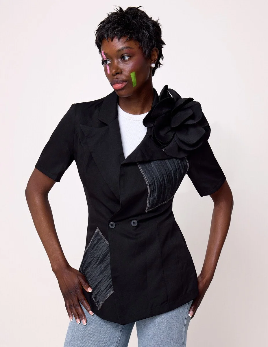JUS10H Black Summer Jacket editorial, three-quarter view of female model in black double-breasted blazer with oversized floral corsage and multiple gray denim patchwork panels, white tee, light-wash jeans, white background, photographed by Scott Park