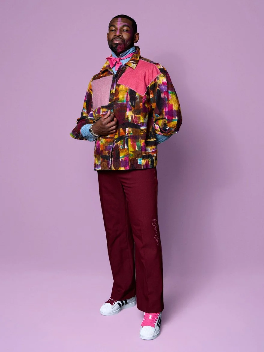 A man standing against a purple background, wearing a colorful patchwork jacket, maroon pants, and pink and white sneakers, with a pink bandana around his neck and a face paint design on his forehead.