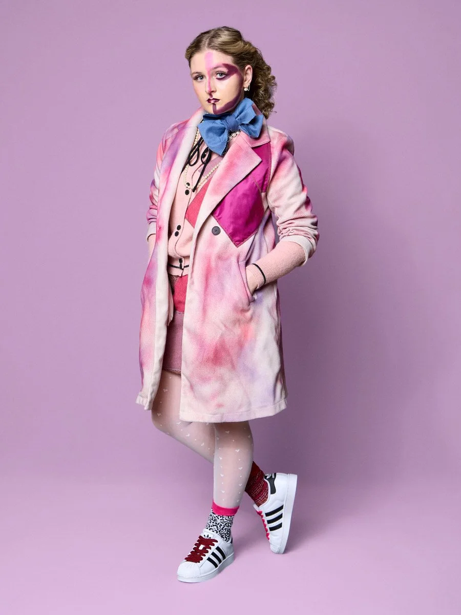 A young woman with colorful, artistic makeup and curly hair, wearing a pastel pink coat over a pink outfit, with a blue bow-tie, patterned socks, and sneakers, standing against a lavender background.