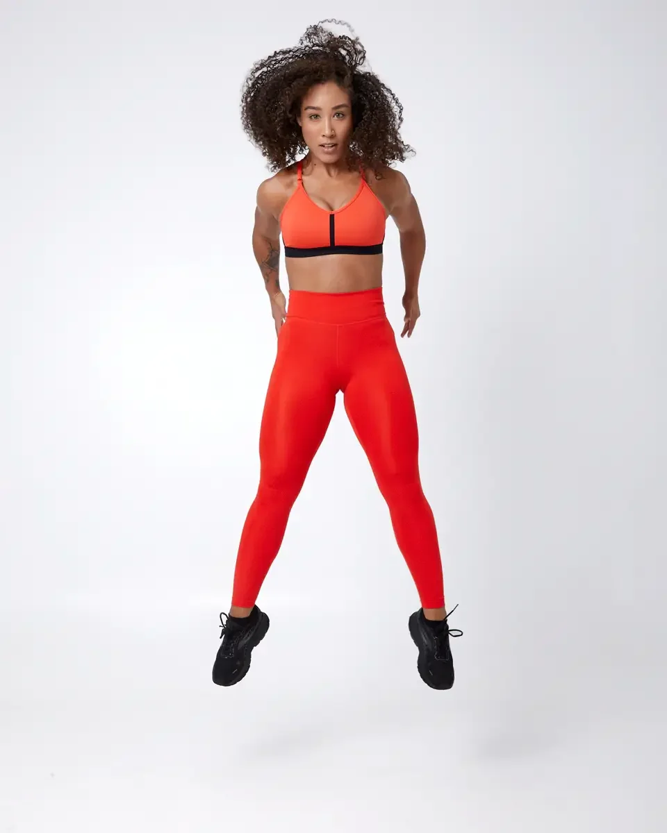 Model portfolio development session, commercial athletic look, orange sports bra with black underband, high-waist orange leggings, black athletic sneakers, full-length, mid-jump, direct gaze, white background