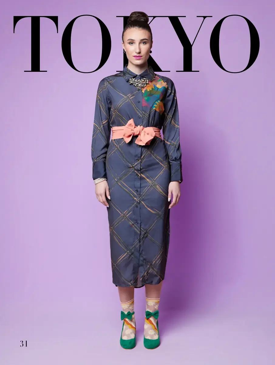 JUS10H 2025 Tokyo lookbook, a model wearing a slate blue diagonal plaid button-down midi dress with a colorful floral appliqué at the chest, a coral satin bow belt, pearl bracelet and collar necklace, daisy-print sheer socks, and green platform Mary 