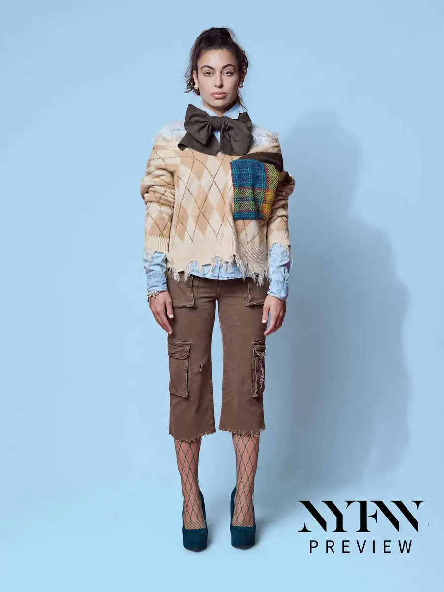 JUS10H 2025 social media, model in camel argyle fringe sweater with multicolor tartan patchwork panel, olive oversized bow, and brown distressed cargo capris over fishnet tights and teal platform heels, light blue background, photographed by Scott Pa