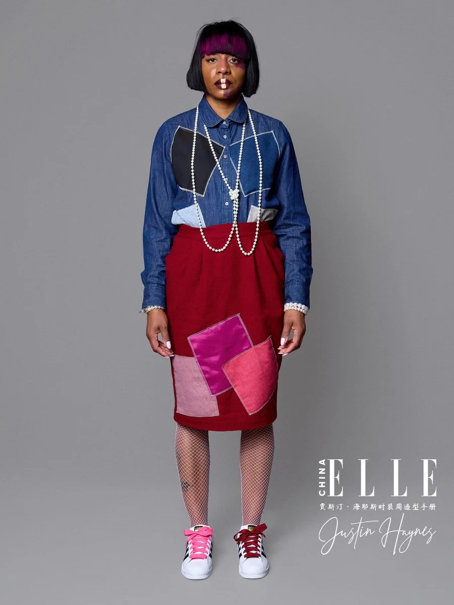 A woman with black and purple hair wearing a denim shirt with colorful patches, red skirt with pink and purple fabric squares, fishnet stockings, white sneakers with pink shoelaces, and layered pearl necklaces, standing against a gray background.