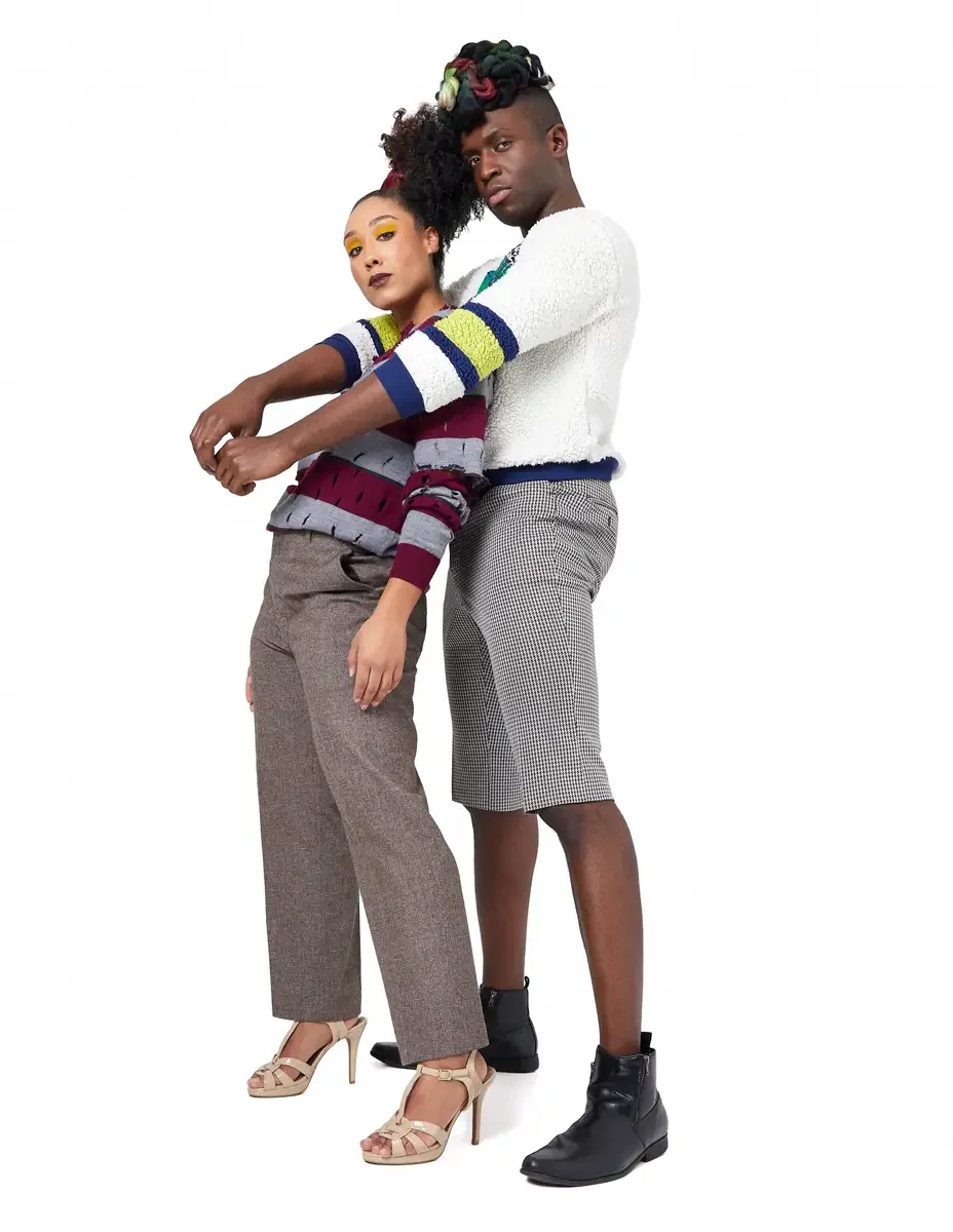 Model portfolio development session, high fashion editorial look, two models in front-to-back stance, woman in burgundy and heather grey destroyed-knit stripe sweater and straight-leg heathered trousers, man in white patchwork bouclé knit sweater and