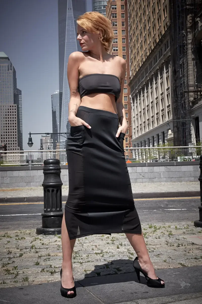 Satine, full length, three-quarter turn, hands in pockets, black strapless bandeau crop top, black satin fitted midi skirt with wide waistband and front slit hem, black satin peep-toe stiletto heels, Lower Manhattan street, One World Trade Center in 