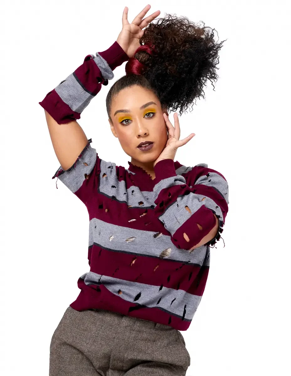 Model portfolio development session, high fashion editorial look, burgundy and heather grey destroyed-knit stripe sweater with raw-edge cutout sleeves, blazer removed, three-quarter, one arm raised overhead, one hand at face, dynamic pose, direct gaz