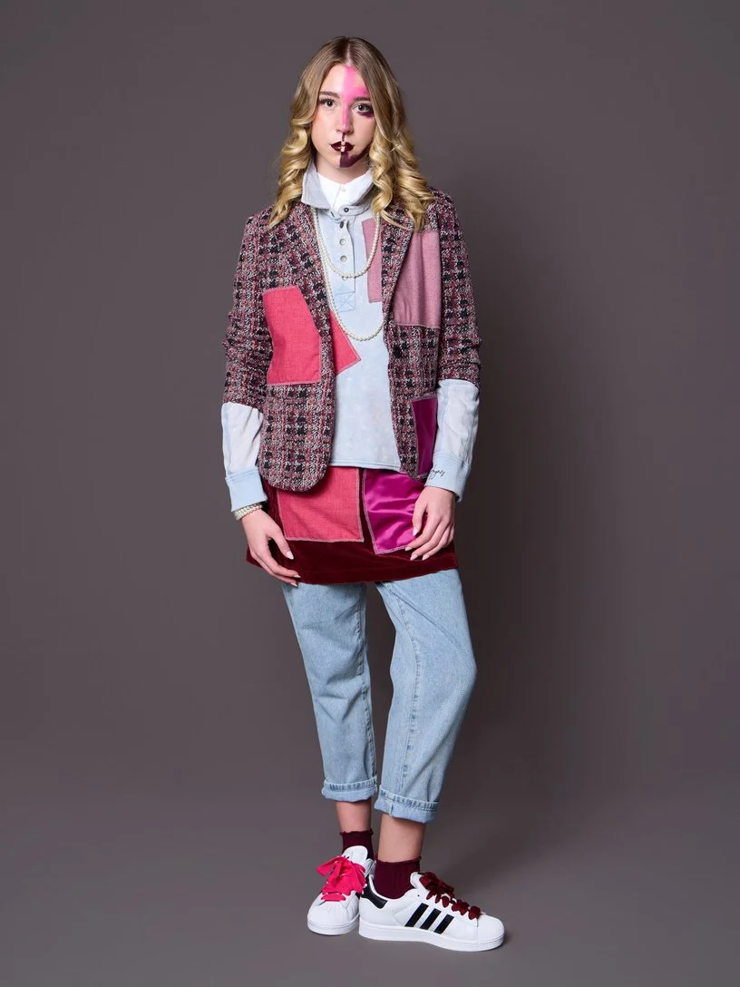 Young woman in a colorful patchwork blazer, pastel shirt, pink and burgundy skirt, light-wash jeans, and white sneakers with red laces, standing against a gray background.