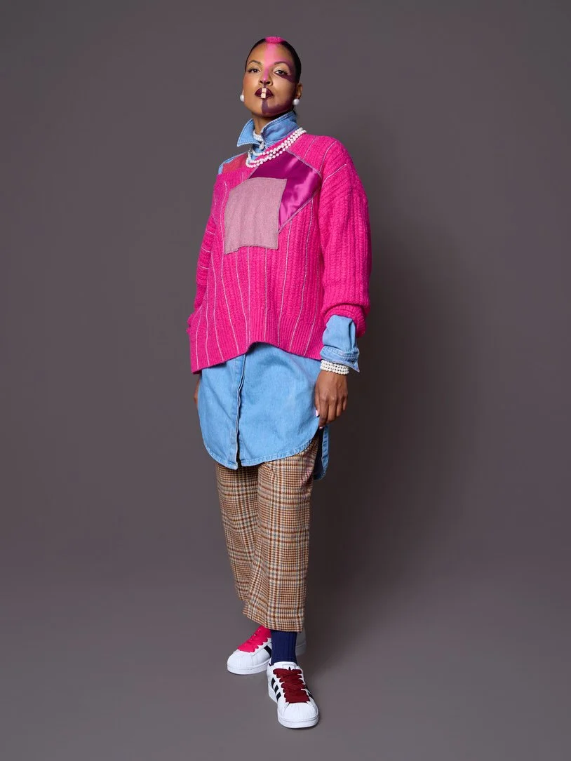 A woman wearing a pink sweater layered over a blue shirt, paired with plaid pants and sneakers, standing against a gray background.
