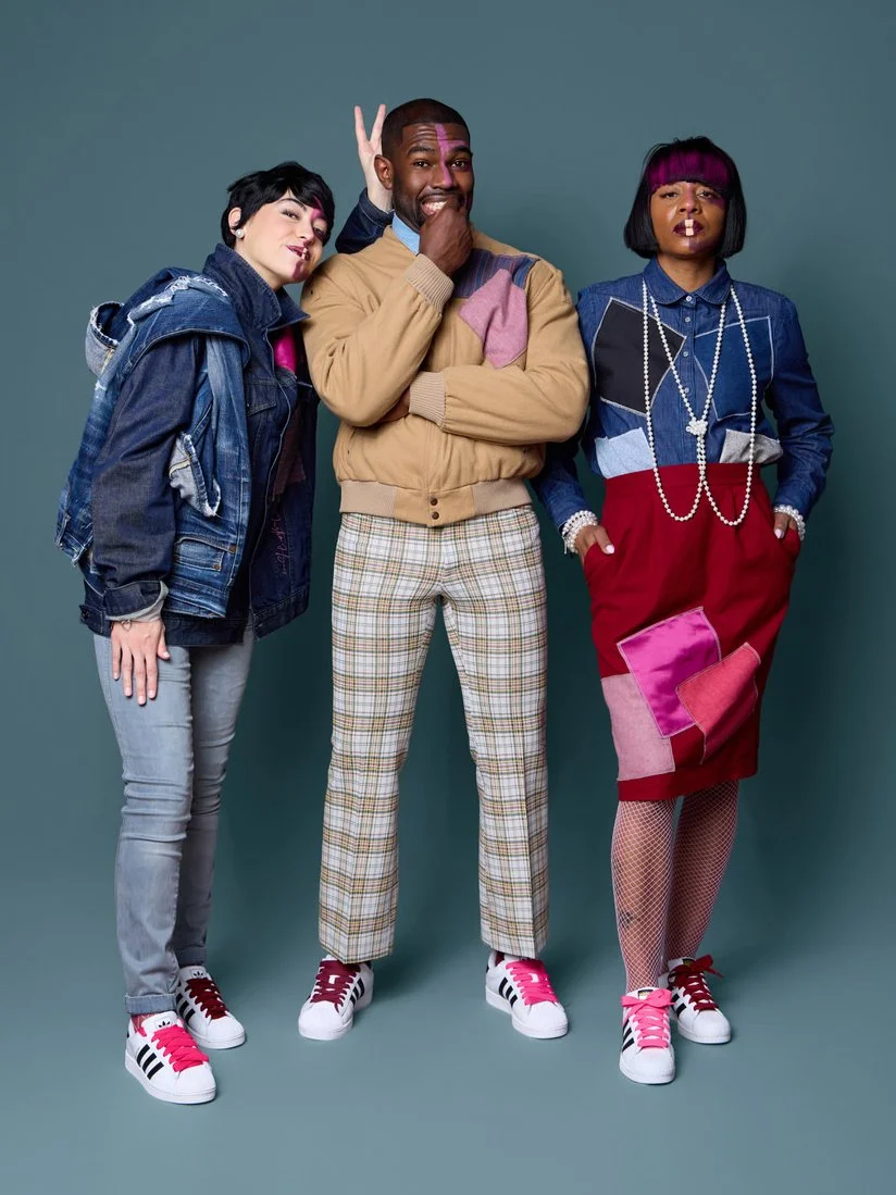 Three people standing together against a solid background. The person on the left is wearing a denim jacket, gray pants, and white sneakers with pink and black accents. The person in the middle is wearing a beige sweater with color blocks, plaid pant
