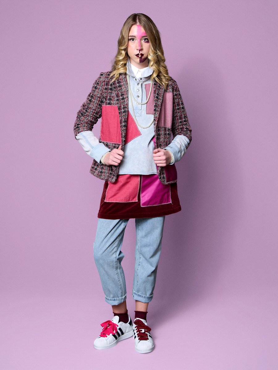 Young woman with blonde wavy hair stands against a purple background, wearing a colorful patchwork jacket, layered shirts, a skirt, light blue jeans, and sneakers with pink and black laces.