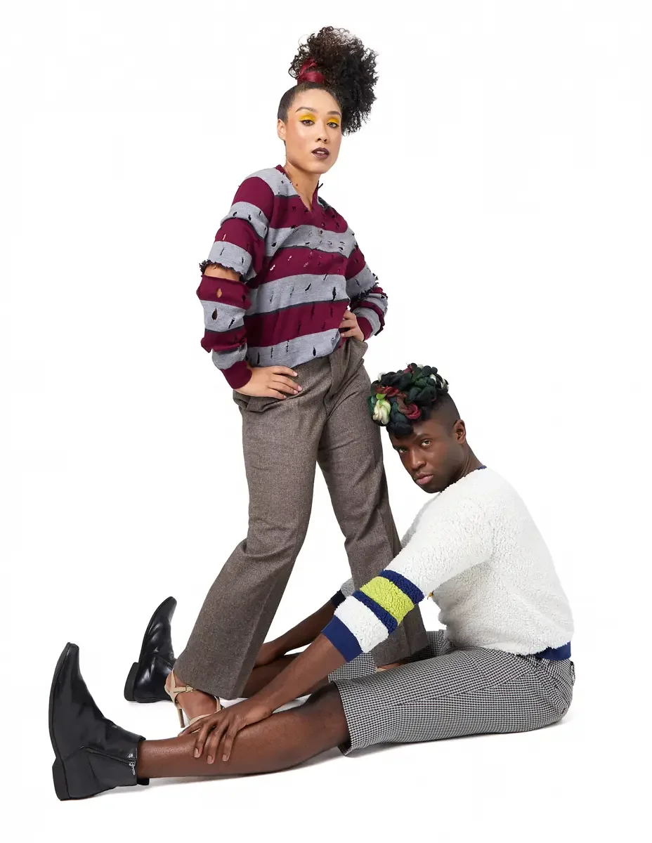 Model portfolio development session, high fashion editorial look, woman standing in burgundy and heather grey destroyed-knit stripe sweater and straight-leg heathered trousers with hand on hip, man seated on floor in white patchwork bouclé knit sweat