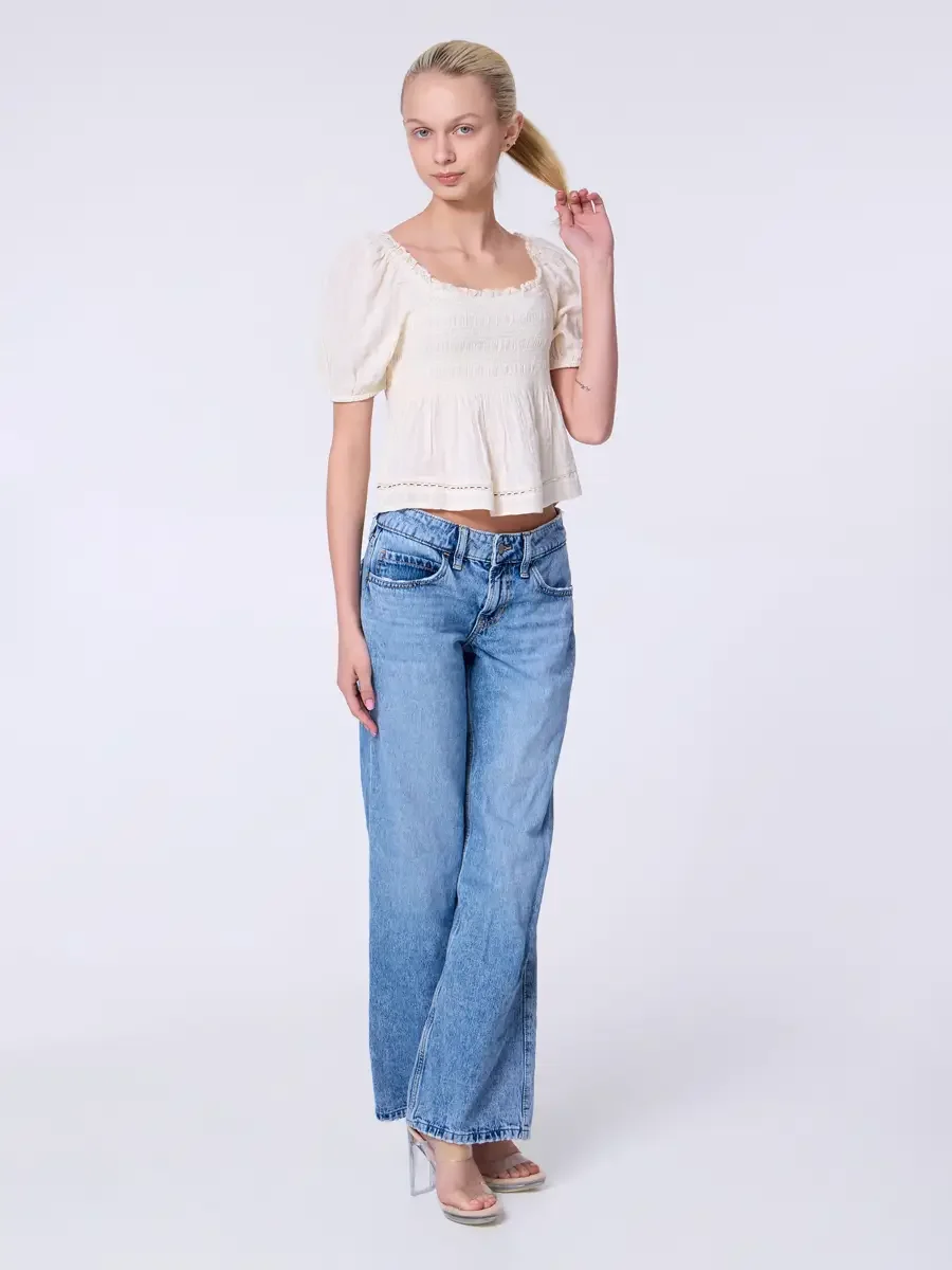 Model portfolio development session, commercial look, cream smocked square-neck puff-sleeve crop top and low-rise light wash wide-leg jeans, full-length, slight turn, one hand raised to ponytail, direct gaze, smocked bodice and crochet hem trim fully