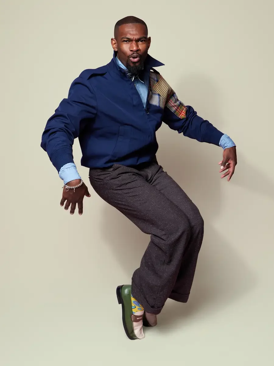 JUS10H 2025 Billboard Magazine UAE feature, model mid-movement in navy harrington jacket with multicolor patchwork sleeve panel over chambray shirt and wide-leg charcoal trousers, sage background, photographed by Scott Parker Photo