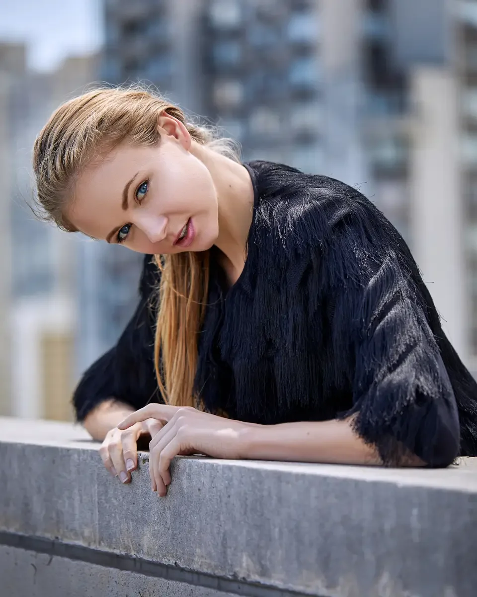 Julia, close-up portrait, leaning on the rooftop railing and turned toward camera, black tiered fringe jacket with layered construction over black fitted dress, hair slicked back in a low ponytail, Jersey City rooftop test shoot, photographed by Scot