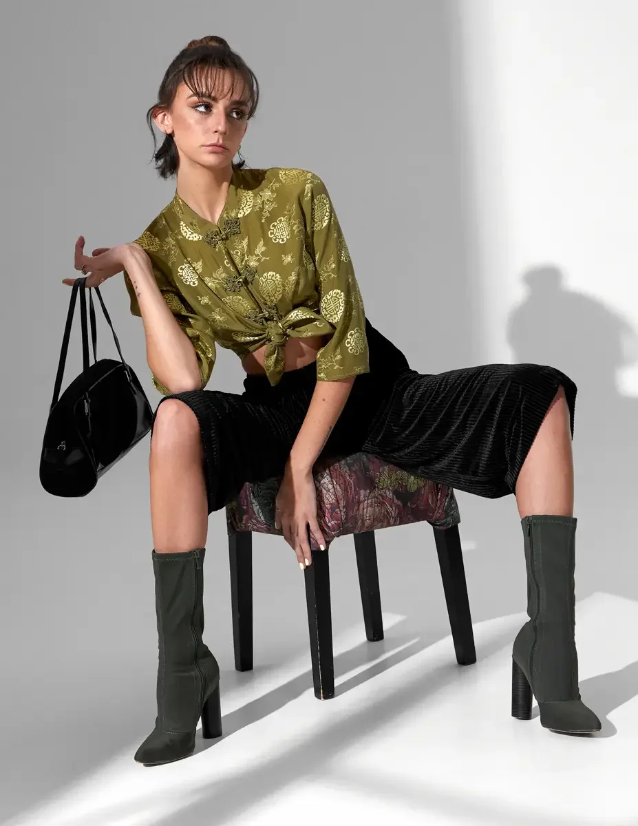 Macy, full length, seated on upholstered stool, olive green satin jacquard tied crop top with gold medallion print and frog closures, black wide-leg ribbed velvet culottes, olive green block-heel sock boots, black patent barrel bag, New York studio t